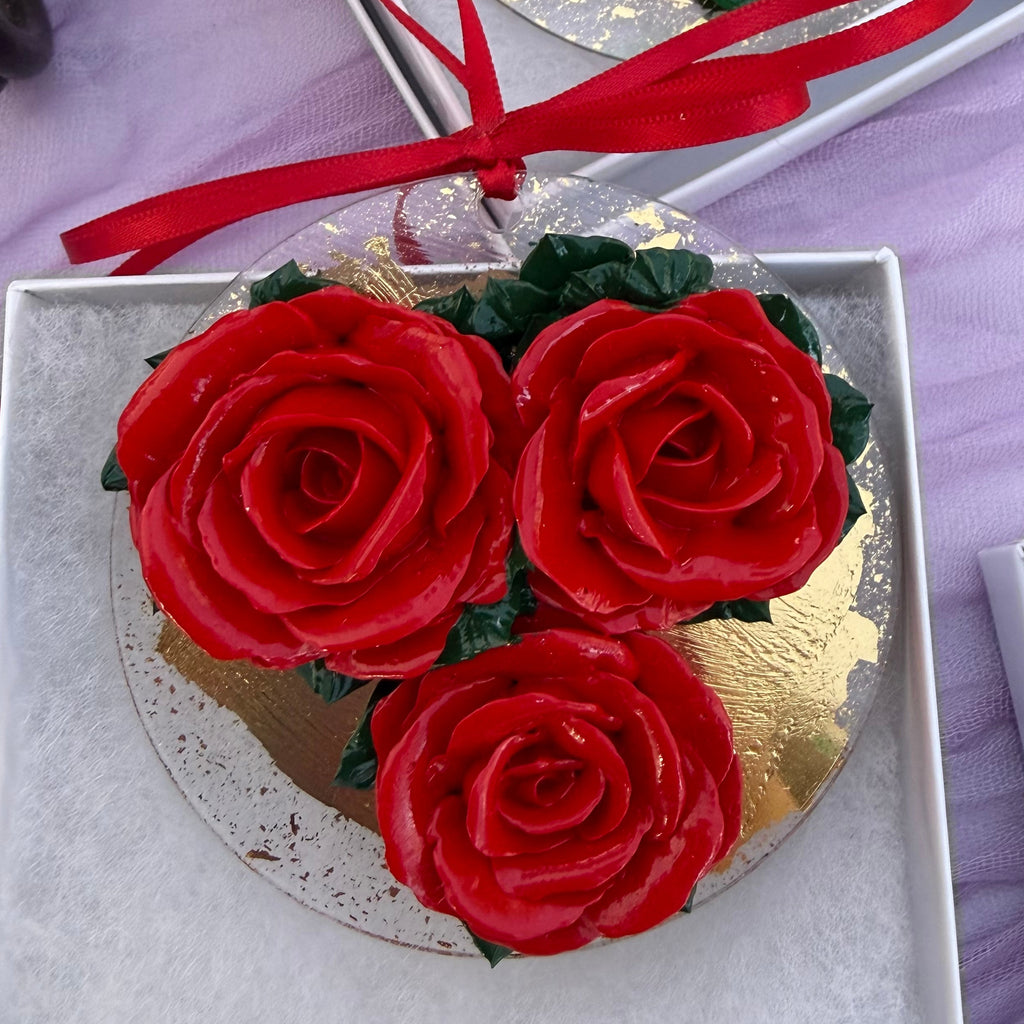 Three red rose-shaped ornament with green leaves and gold circular bases, with red ribbon